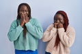 Two african woman standing over blue background laughing and embarrassed giggle covering mouth with hands, gossip and scandal Royalty Free Stock Photo