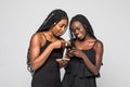 Two african women expressing excitement or surprise while both using cell phone isolated on gray background