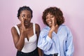 Two african women standing over pink background hand on mouth telling secret rumor, whispering malicious talk conversation Royalty Free Stock Photo