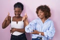 Two african women standing over pink background looking proud, smiling doing thumbs up gesture to the side Royalty Free Stock Photo