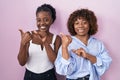 Two african women standing over pink background pointing to the back behind with hand and thumbs up, smiling confident Royalty Free Stock Photo