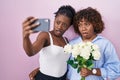 Two african women taking a selfie photo with flowers in shock face, looking skeptical and sarcastic, surprised with open mouth