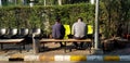 Two Asian man sitting on wooden bench among sunlight in the morning beside steel pole with ashtray garbage and trash can Royalty Free Stock Photo