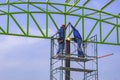 Two Asian welders on scaffolding are welding metal roof structure of industrial building against blue sky