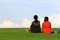 Two Asian women in black and red dress sitting on grass field and relaxing on weekend at garden park with cloud and blue sky