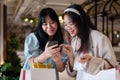 Two Asian women chat happily in a shopping mall corridor, looking at a smartphone together Royalty Free Stock Photo