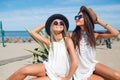 Two attractive brunette and blonde girls with long hair are sitting on the beach near sea. They wear hats, sunglasses Royalty Free Stock Photo