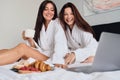 Two attractive cheerful women in white bathrobes joyfully watching movie on laptop together with delicious pastry and