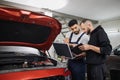 Two attractive mechanics men standing and looking at laptop with tablet and under car hood, Royalty Free Stock Photo