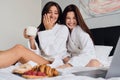 Two attractive women in white bathrobes happily watching movie on laptop together with delicious pastry and coffee