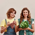 Two attractive young girls, twin sisters in casual wear holding fresh green salad and piece of meat isolated over light