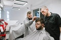 Two barbers are cutting a bearded man in a light modern barber shop. Hairdresser teacher teaches student to do hairstyles. Barber