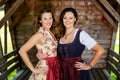 Two bavarian women in dirndl standing by a wooden hut
