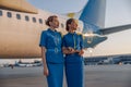 Two beautiful air hostesses in blue uniform smiling away, standing in front of a big passenger airplane in airport at Royalty Free Stock Photo