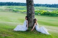 Two beautiful brides sitting near the tree on the green field