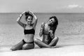 Two beautiful cheerful young women twin sisters in bikini sitting on sand on the beach at sunny day Royalty Free Stock Photo