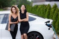Two beautiful girls in black outfut smiling and posing. Businesswomen with laptop and electric car with charging socket plugged in