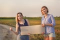 Two beautiful girls get hitchhiked and vote with a sign on cars on a rural road in the rays of sunset.