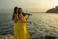 Two beautiful girls violinists in yellow concert dresses are playing electric violins with sunset, mountains and Mediterranean sea