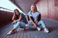 Two beautiful happy hipster girls sitting on skateboard at a sidewalk under bridge. Royalty Free Stock Photo