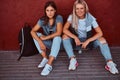 Two beautiful hipster girls sitting on a skateboard and leaning on a wall. Royalty Free Stock Photo