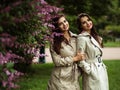 Two beautiful twins young women in trench coats near blooming lilac