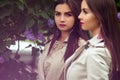 Two beautiful twins young women in trench coats near blooming lilac