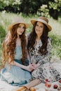 Two beautiful friend womens in summer dresses and straw hats enjoying picnic in countryside. Royalty Free Stock Photo