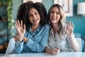 Two beautiful women waving hand while doing video call looking at camera in the office at home Royalty Free Stock Photo