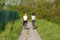 Two beautiful young girls in sport outfit riding their bicycles on paved country path surrounded with high green grass and forest