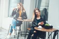 Two beautiful young women in fashionable wears sitting outdoor in cafe and using smartphones while drinking coffee. Business messa Royalty Free Stock Photo