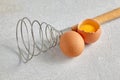 Two beige hen's eggs with yolks and a spiral whisk to whip on a gray concrete countertop