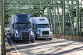Two big rig semi trucks blue and white transporting cargo in dry van semi trailers running side by side on the truss arched bridge