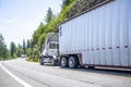 Two big rigs semi trucks with different semi trailers running towards each other on the winding mountain road with cliff and trees