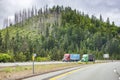 Two big rigs semi trucks transporting cargo in semi trailers driving side by side on the highway road along the forested mountain Royalty Free Stock Photo