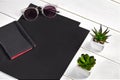 Two black sheets of paper, notebook, sunglasses and green succulents in pots on white wooden tabletop or background