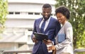 Two black smiling coworkers checking financial reports outdoors, woman having coffee