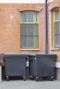 Two black waste trash containers under the windows of a brick building