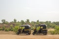 A two black and yellow Indian motor rickshaws stand on an asphalt road against the background of a hill and green forest