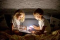 Two blond cute childrem, boy and girl, siblings, lying under the cover in bed, reading book together with small light Royalty Free Stock Photo