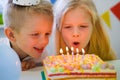 Two blonde caucasian kids boy and girl have fun blowing out candles at birthday rainbow cake with burning candles at Royalty Free Stock Photo