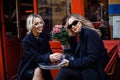 Two blondes in black sit at cafe table in Paris. Young woman smile while looking at lady with red lips and sunglasses. Royalty Free Stock Photo