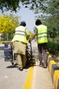 Two blue-collar workers wearing safety jackets are operating a paint-machine to paint draw road markings.