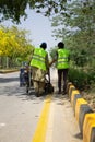 Two blue-collar workers wearing safety jackets are operating a paint-machine to paint draw road markings.