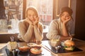 Two bored young women sit at table and look at food. They keep hands under chin. Models are in cafe. Sun is shining