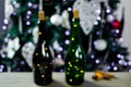 Two bottles of champagne with a glowing garland stand on a wooden table against the background of the Christmas tree. Defocused