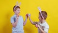 Two boys with rabbit ears on a yellow background beat Easter eggs. Easter
