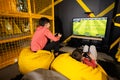 Two brothers playing football video game console, sitting on yellow pouf in kids play center Royalty Free Stock Photo