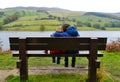 Two brothers sitting on bench with older brother cuddling younger sibling. Overlooking river and hills.