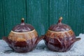 Two brown ceramic pots covered with lids on a table in white snow against a green wooden wall Royalty Free Stock Photo